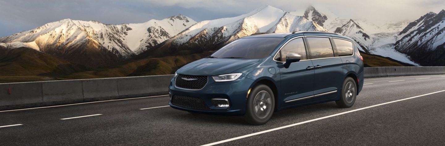 A blue 2023 Chrysler Pacifica Pinnacle Hybrid being driven on a highway with snow-capped mountains in the distance.