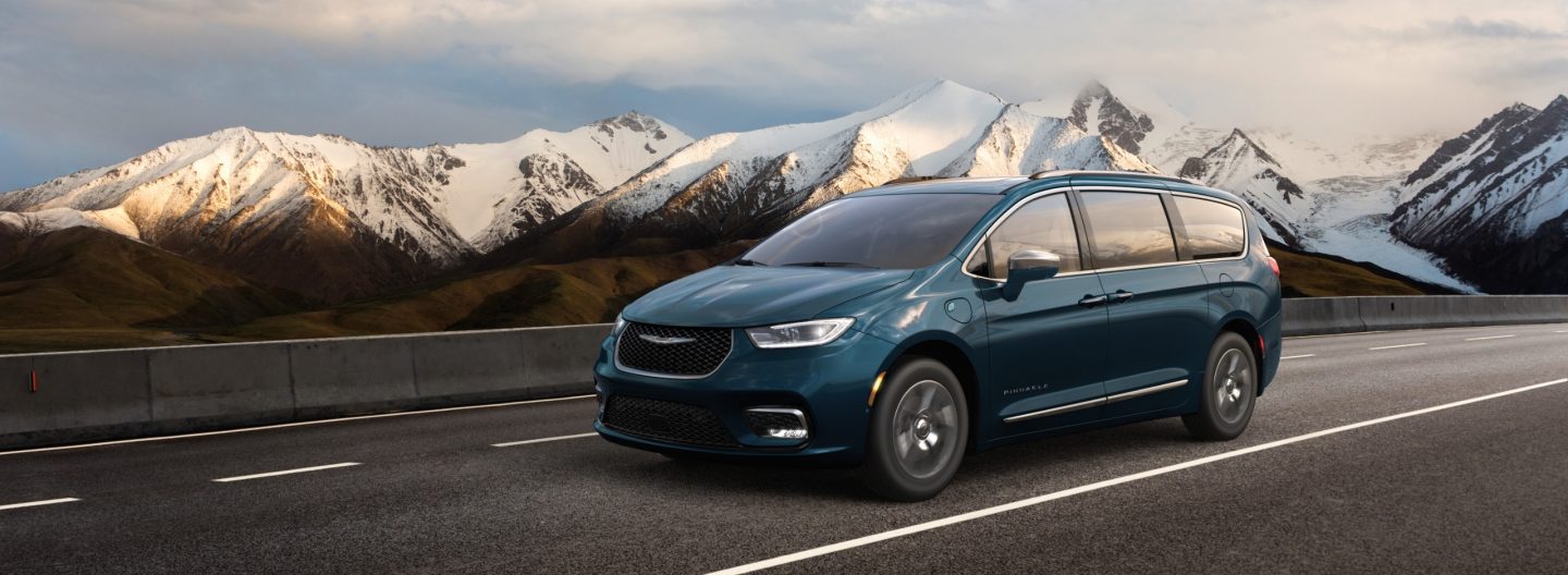 A blue 2024 Chrysler Pacifica Pinnacle Plug-In Hybrid being driven down a highway with snow-capped mountains in the background.