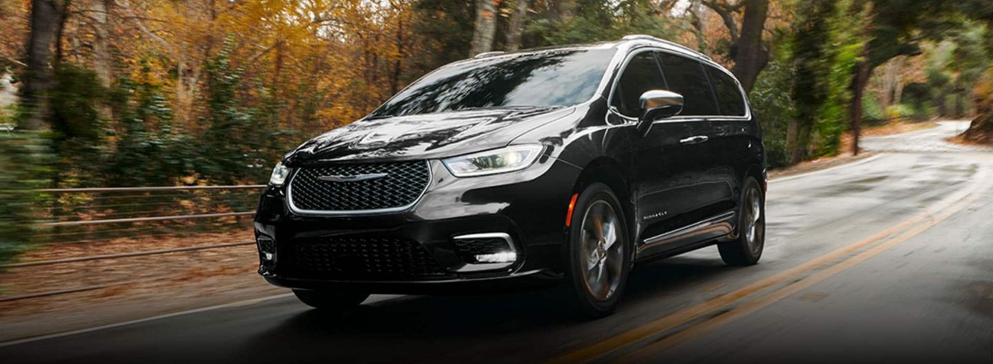 An angled front profile of a black 2024 Chrysler Pacifica Pinnacle being driven on a rural highway. The background is blurred to indicate the vehicle is in motion.