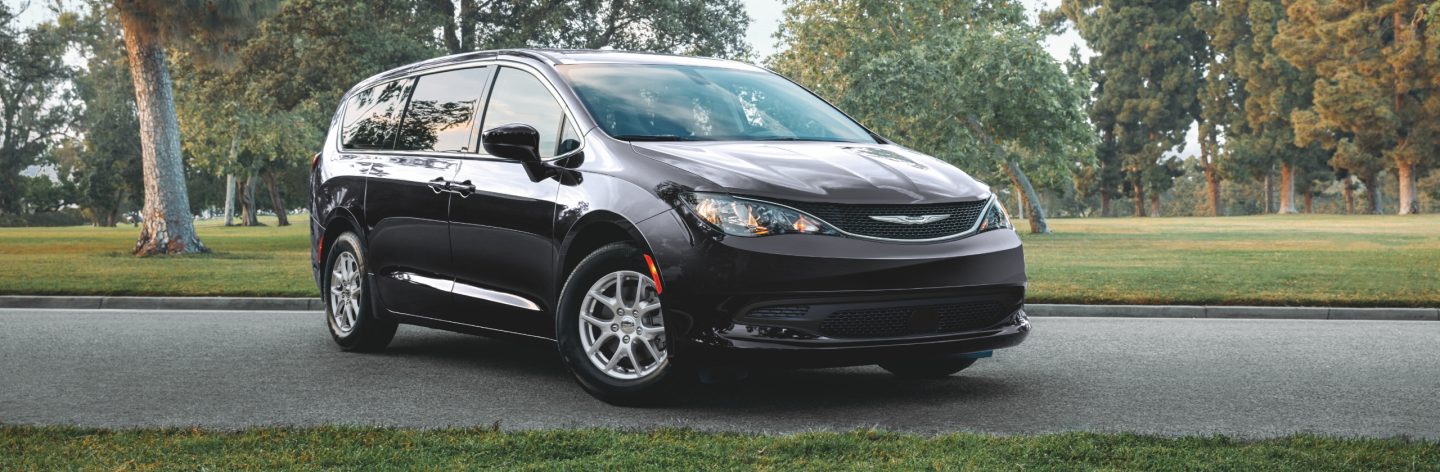 An angled front passenger-side profile of a black 2024 Chrysler Voyager LX parked beside a park.