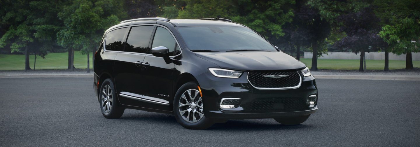 A passenger-side front angle of a black 2026 Chrysler Pacifica Pinnacle Plug-in Hybrid parked on a lot with trees in the background.
