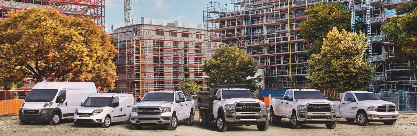 A lineup of six 2022 Ram trucks and vans in front of a tall building under construction, covered in scaffolding.