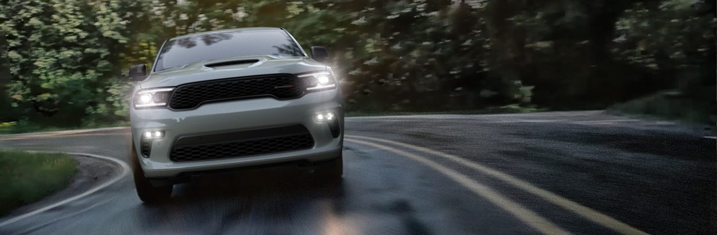 A silver 2023 Dodge Durango R/T with its headlamps on, taking a curve on a wet roadway.