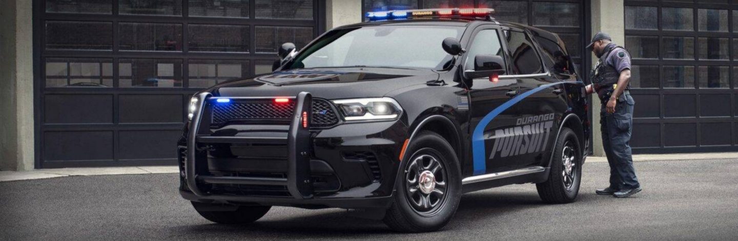 The 2025 Dodge Durango Pursuit with its flasher front end lights and beacon lights on, parked outside an industrial garage with a police officer near the back of the vehicle.