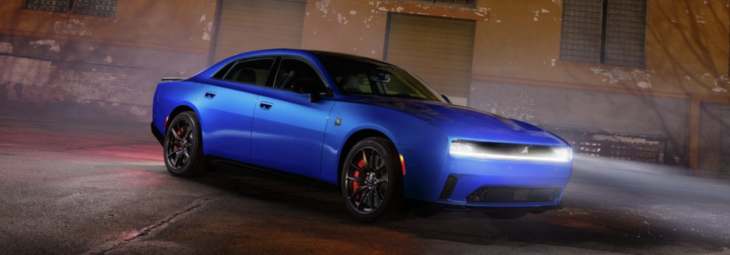 A passenger-side front angle of a blue 2026 Dodge Charger Daytona Scat Pack on a road at night with its headlights on.