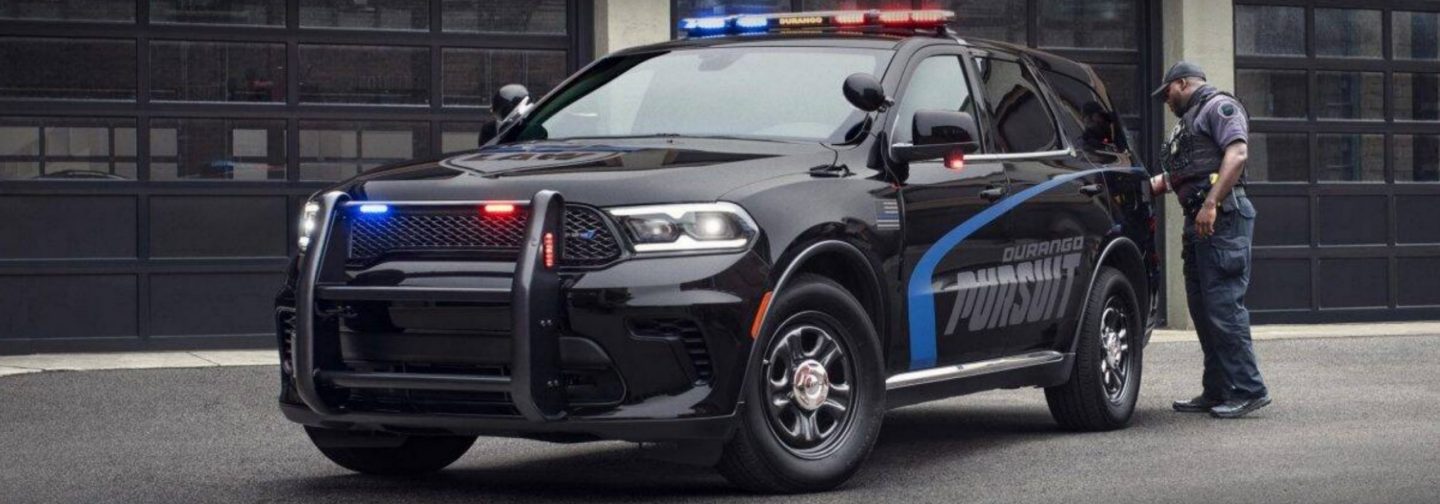 The 2026 Dodge Durango Pursuit with its flasher front end lights and beacon lights on, parked outside an industrial garage with a police officer near the back of the vehicle.
