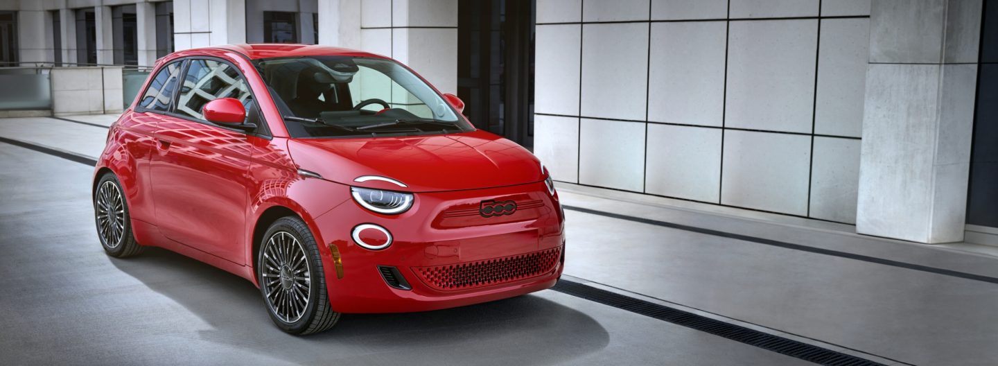 An angled front passenger-side profile of a red 2024 Fiat (Red) 500e Edition parked beside a contemporary building.