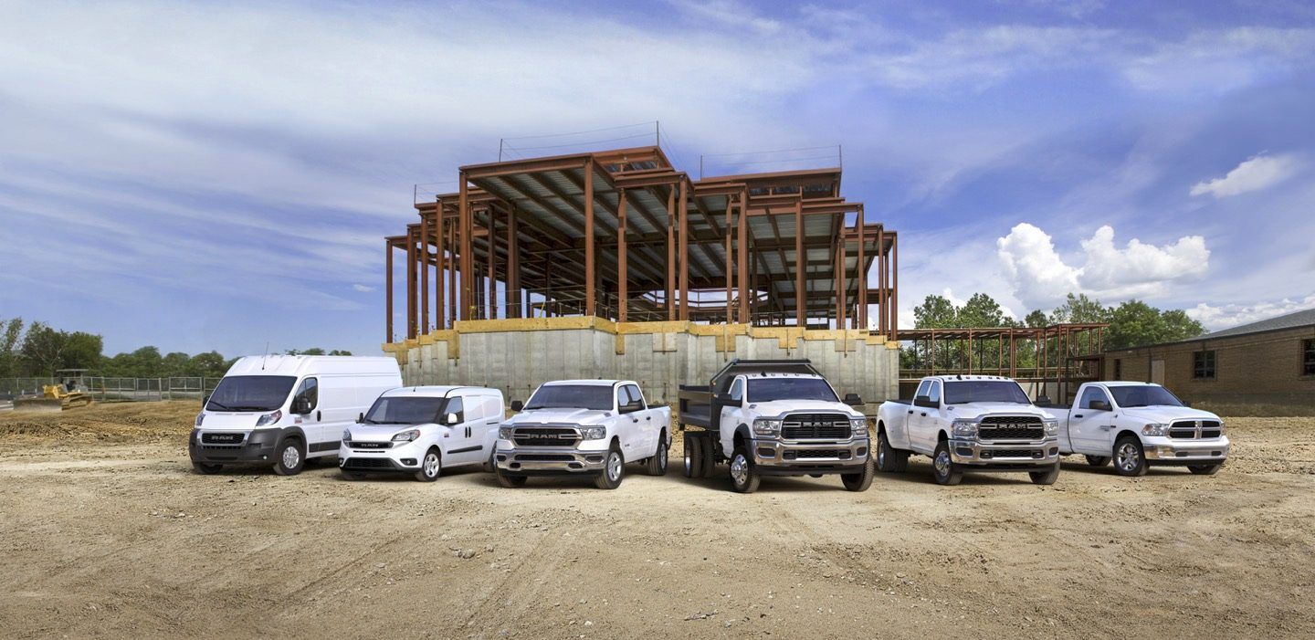 A lineup of six Ram trucks and vans, parked on a dirt lot in front of a framed-in building.