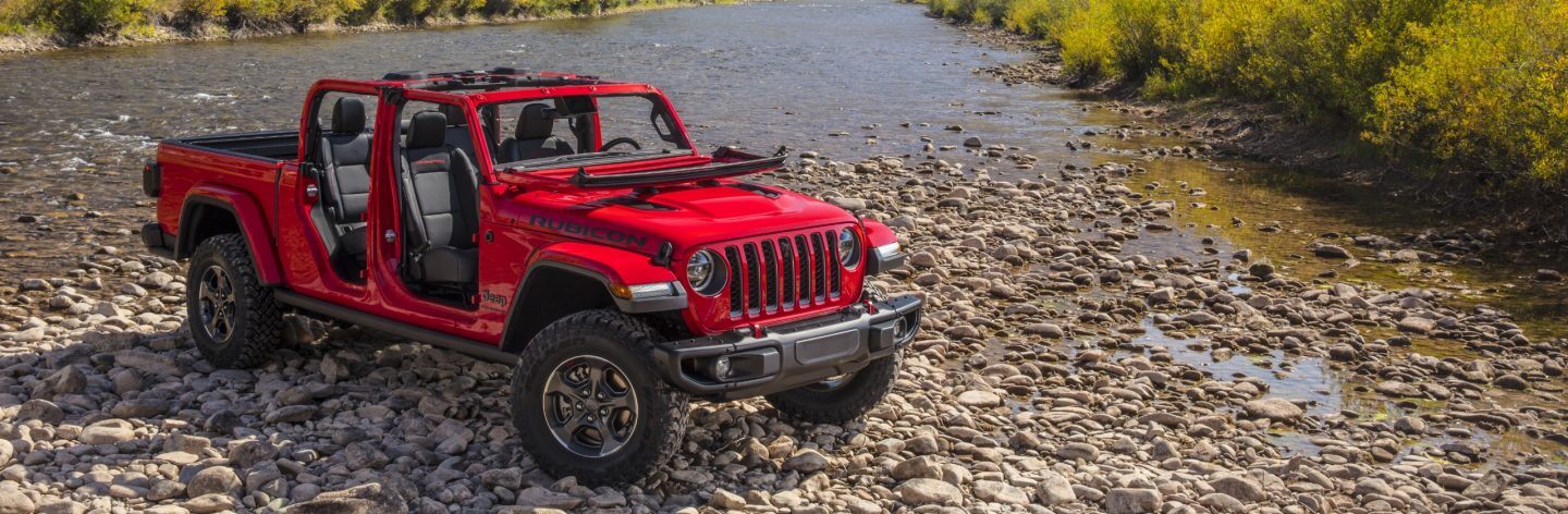 A red 2023 Jeep Gladiator Rubicon with its doors and top off and windshield folded down, being driven over a rocky stream.