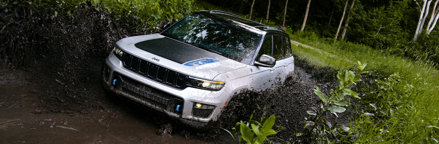 The 2023 Jeep Grand Cherokee Trailhawk 4xe being driven through a mud puddle, splashing mud up to its wheel flares.