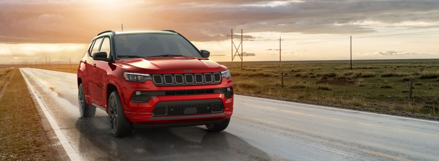 A red 2024 Jeep Compass Limited being driven on a highway between two open fields at sunset.