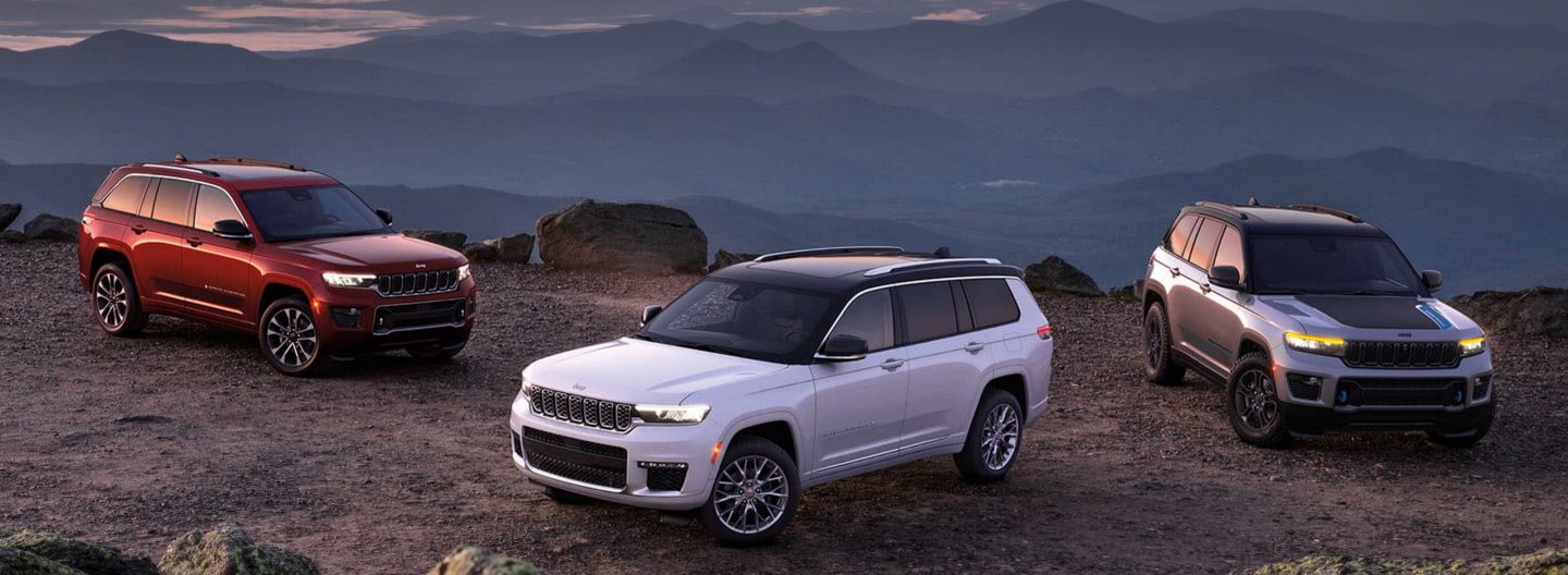 Three 2024 Jeep Grand Cherokee models parked on a gravel mountain road at sunset, from left to right: a red Grand Cherokee Overland, a white Grand Cherokee Summit with black roof and a silver Grand Cherokee Trailhawk with black hood insert. 