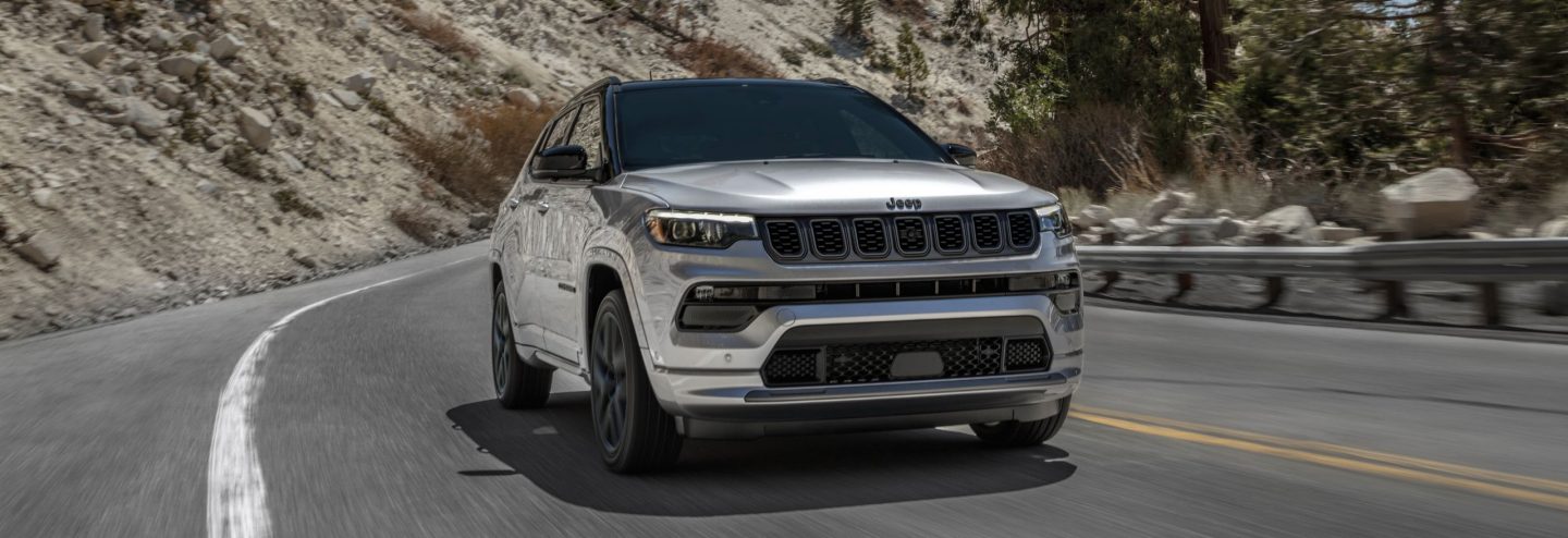 A front angle of a silver 2025 Jeep Compass Limited with the High Altitude Package, traveling on a winding mountain highway. 