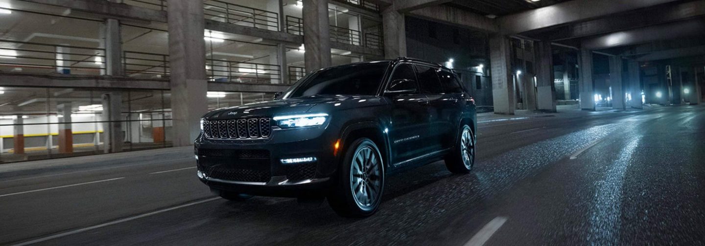 A silver 2025 Jeep Grand Cherokee Trailhawk 4xe with black hood insert, parked on a clearing in the mountains, with a dense forest in the background. 