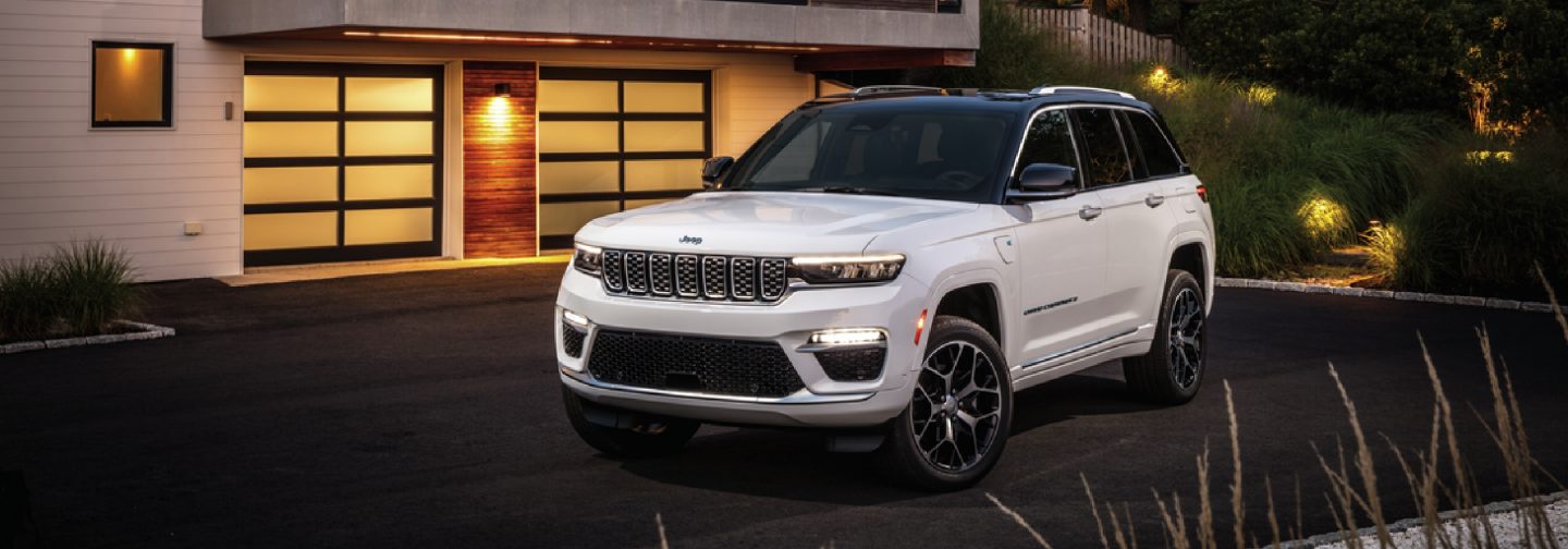 A white 2025 Jeep Grand Cherokee Summit Reserve 4xe parked outside of a contemporary home in the evening. 
