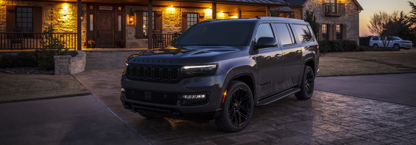 A driver-side front angle of a black 2025 Jeep Wagoneer Carbide, parked in the courtyard of a large, upscale lodge at sunset.