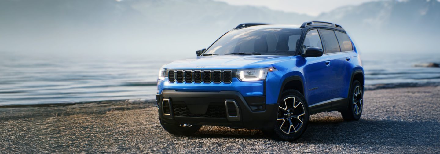 A gray 2026 Jeep Cherokee Overland, traveling on a beach near the water's edge, with mountains in the background.