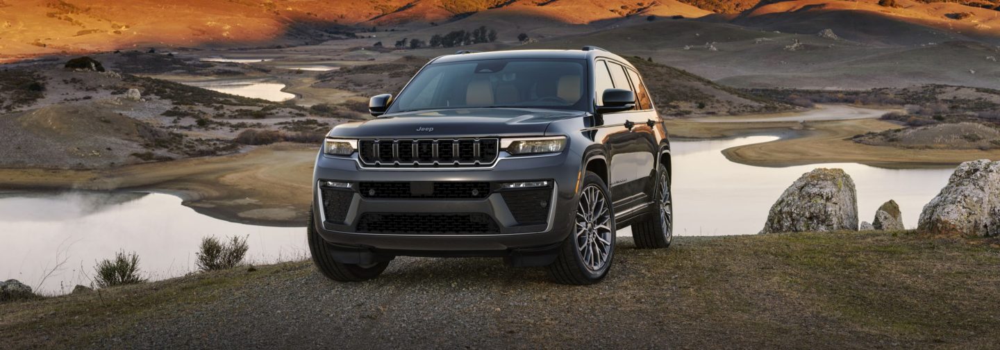 A gray 2026 Jeep Grand Cherokee Summit parked on the edge of an embankment at sunset, with a lake below.