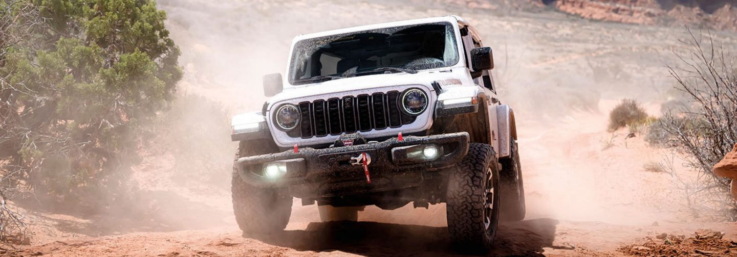 A driver-side profile of a blue gray 2026 Jeep Wrangler Sahara four-door traveling on a city highway.