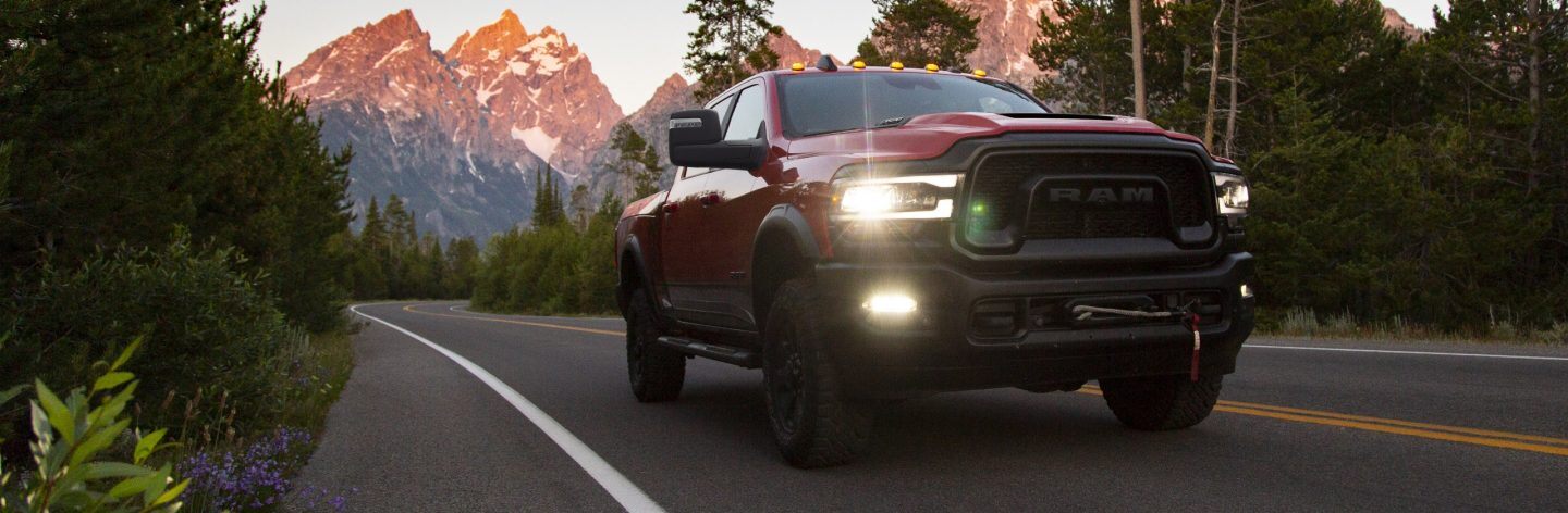 A red 2023 Ram 2500 Rebel Crew Cab with its headlamps on, being driven on a highway with mountains in the background.