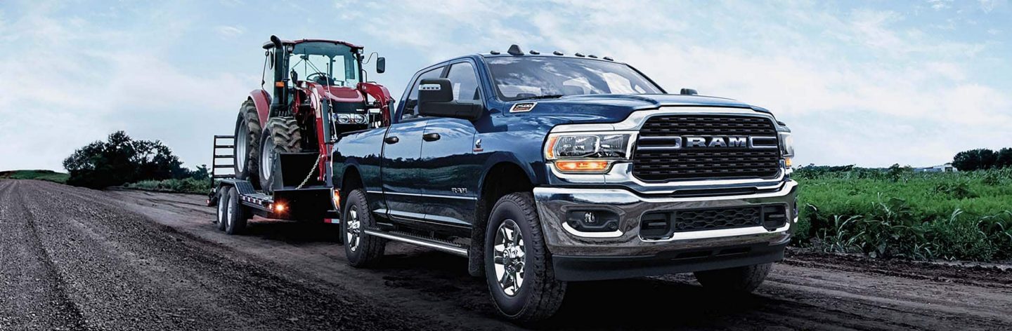 A blue 2024 Ram 2500 Big Horn Crew Cab towing a backhoe on a flatbed trailer as it is driven down a country road.