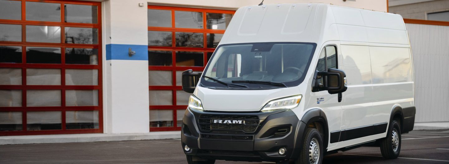 An angled driver-side front profile of a white 2024 Ram ProMaster EV 3500 Super High Roof Delivery Van, parked beside an industrial garage.
