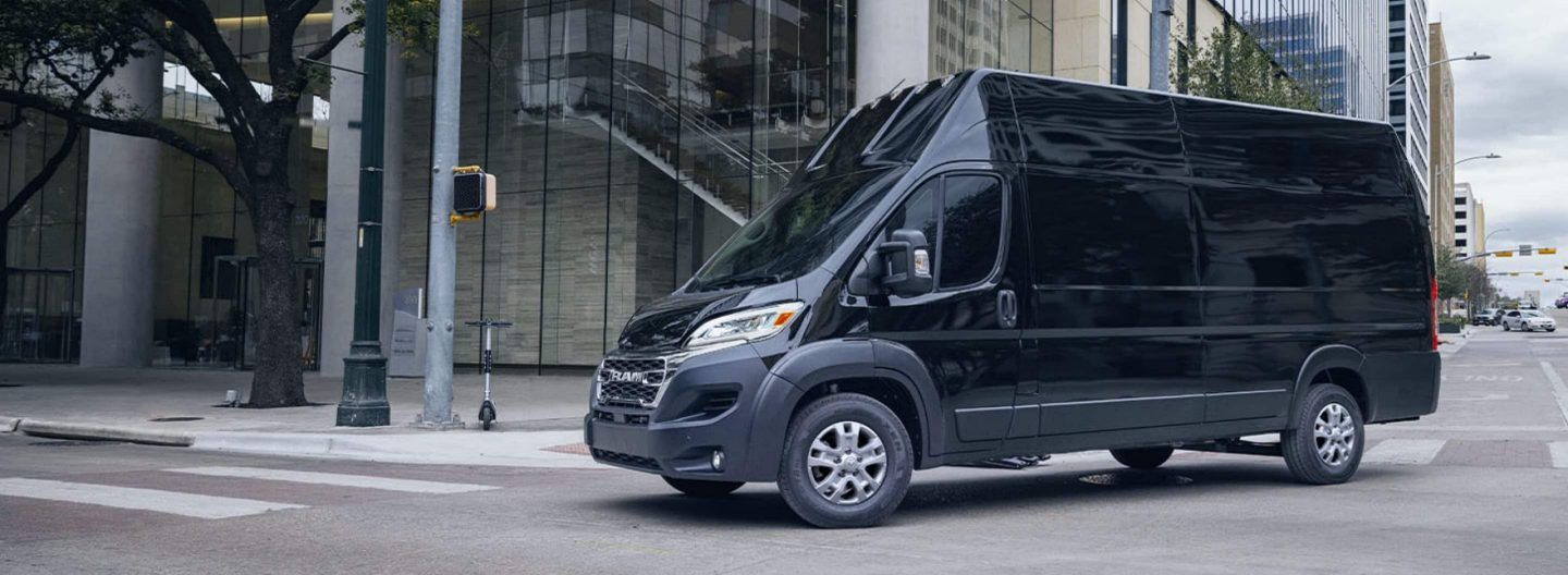 A black 2024 Ram ProMaster 3500 SLT+ Cargo Van Super High Roof making a right turn at a lighted intersection on a city street.