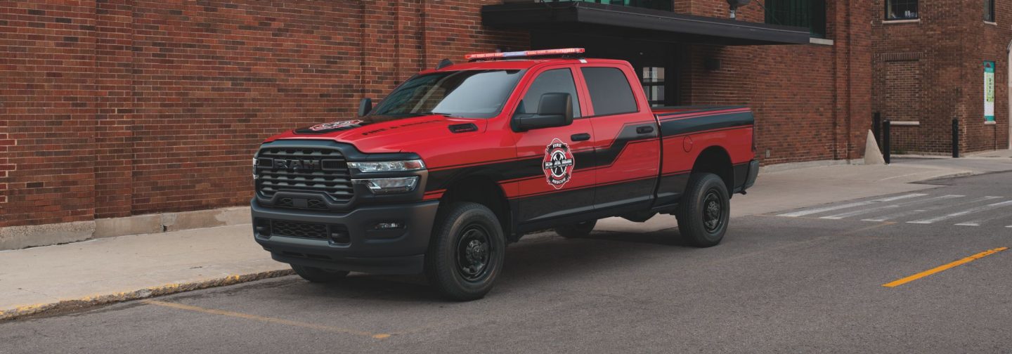 A driver-side front angle of a red 2025 Ram 3500 Tradesman 4x4 Crew Cab with the Special Service Vehicle Group and sample SSV signage on its hood and driver side, parked beside a brick building.