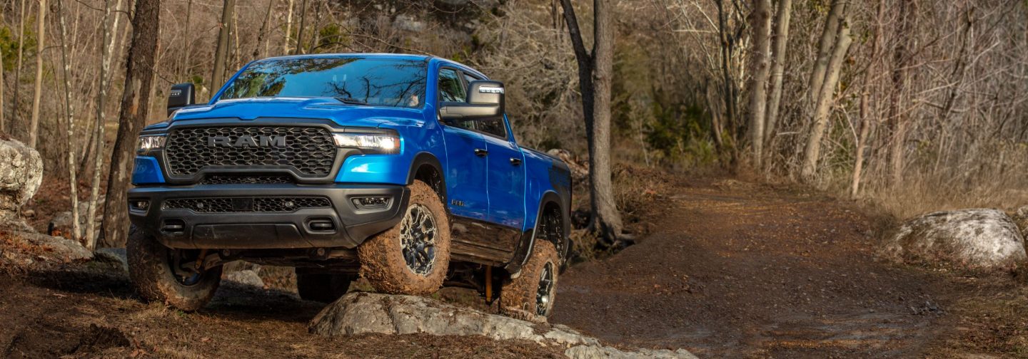 A blue 2026 Ram 1500 Rebel 4x4 Crew Cab ascending a rocky hill, off-road in the woods.