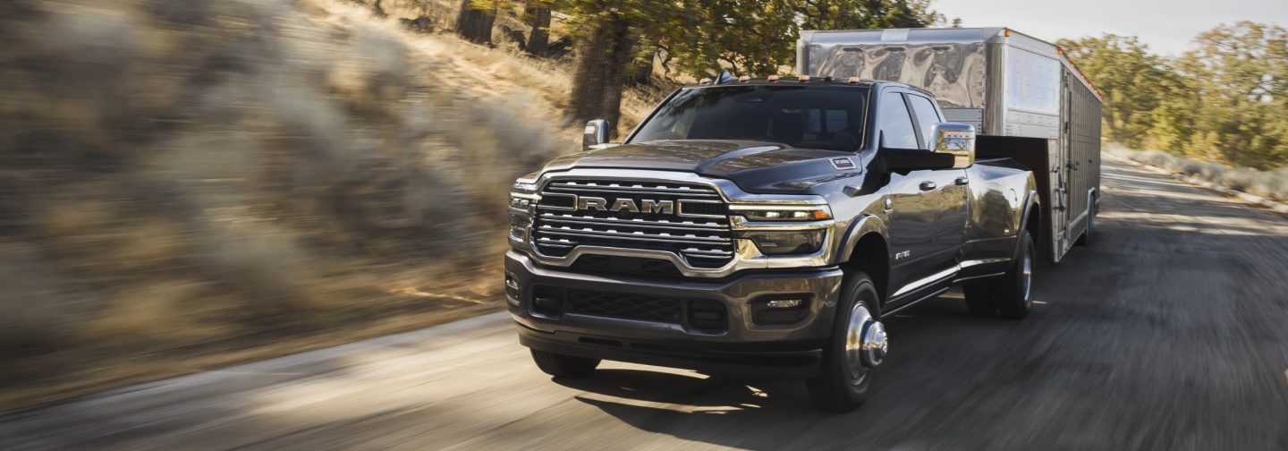 A gray 2026 Ram 3500 Limited Longhorn 4x4 Crew Cab traveling down a highway, towing a large fifth-wheel livestock trailer.