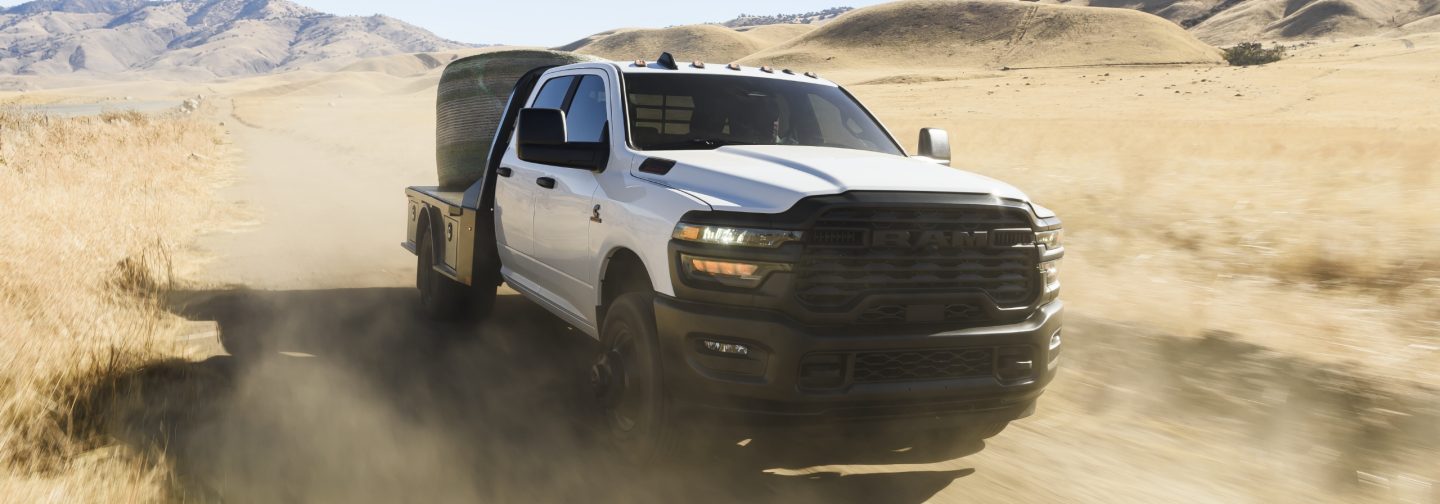 A white 2026 Ram 3500 Tradesman Chassis Crew Cab traveling through a desert with a huge bale of hay on its platform upfit.