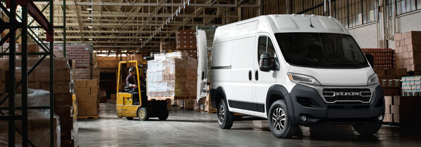 A passenger-side front angle of a white 2026 Ram ProMaster 2500 SLT High Roof Cargo Van, parked in a warehouse with its rear doors fully open and a forklift with a pile of boxes being driven toward the back of the van.