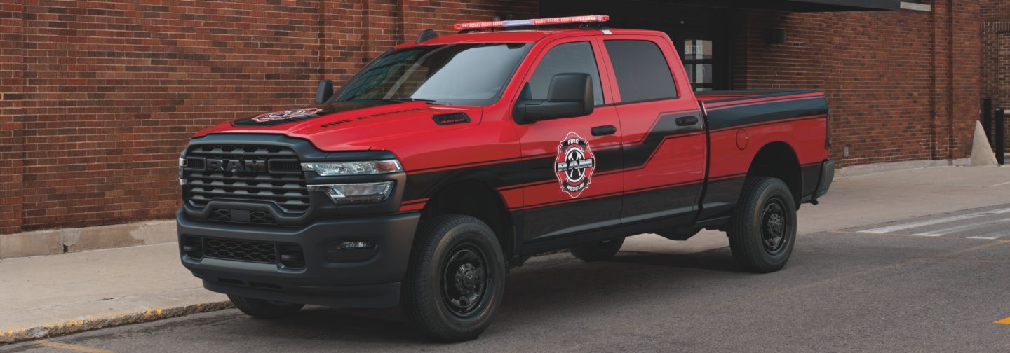 A driver-side front angle of a red 2026 Ram 3500 Tradesman 4x4 Crew Cab with the Special Service Vehicle Group and sample SSV signage on its hood and driver side, parked beside a brick building.