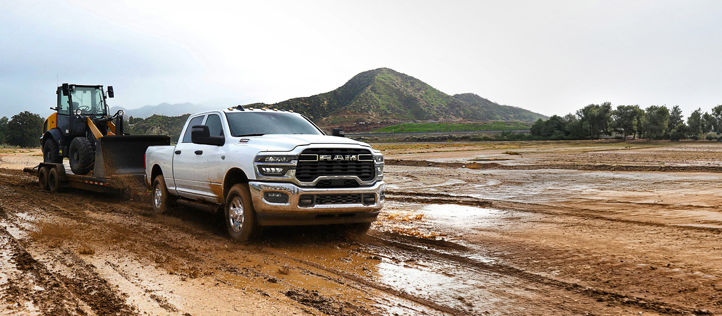 A white 2027 Ram 2500 Heavy Duty Tradesman Crew Cab pickup truck towing a front end loader on a flatbed trailer through a muddy construction site.