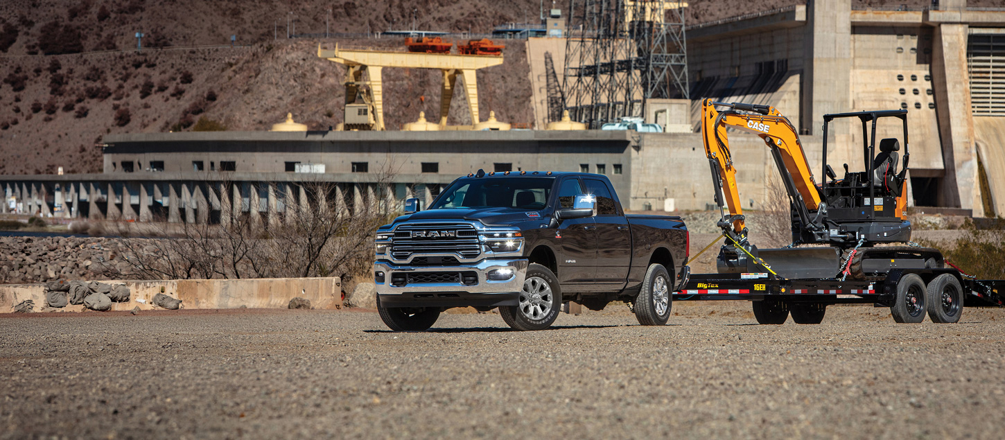 A black 2027 Ram 2500 Heavy Duty Laramie Heavy Duty 4x4 Crew Cab pickup truck towing an excavator on a flatbed trailer, with an industrial complex in the background.