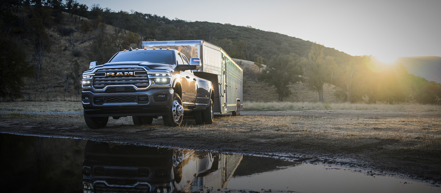 A black 2027 Ram 3500 Heavy Duty Limited Longhorn 4x4 Crew Cab pickup truck towing a large fifth-wheel livestock trailer off-road, beside a body of water.