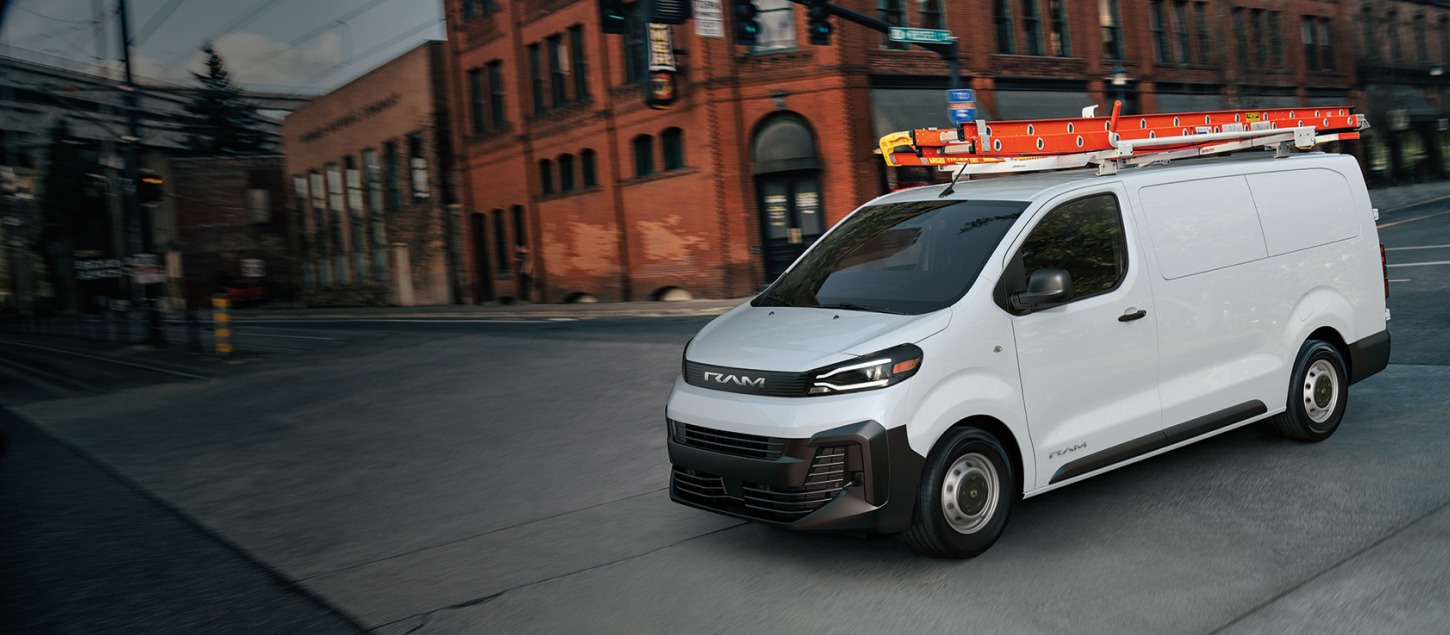 A driver-side front angle of a white 2027 Ram ProMaster City Tradesman Cargo Van with a red ladder on its roof, traveling through an intersection in a city.