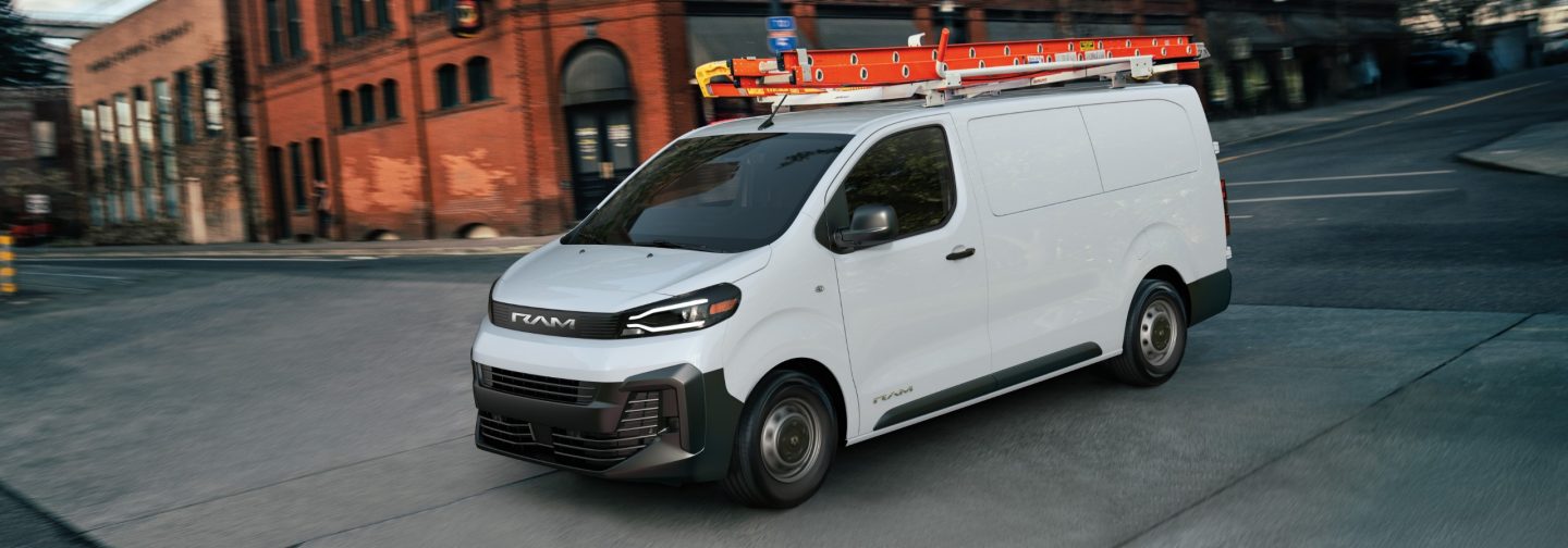 A driver-side front angle of a white 2027 Ram ProMaster City Tradesman Cargo Van traveling through an intersection in a city.